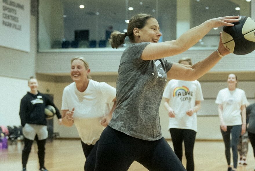 A group of smiling women playing basketball inside a basketball stadium.