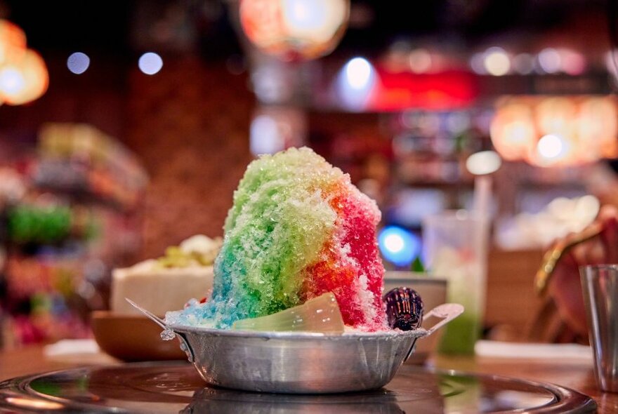 Colorful shaved ice dessert served in a metal dish on a table in a restaurant.