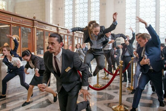 People in a bank wearing corporate attire, leaping, running and dancing. 