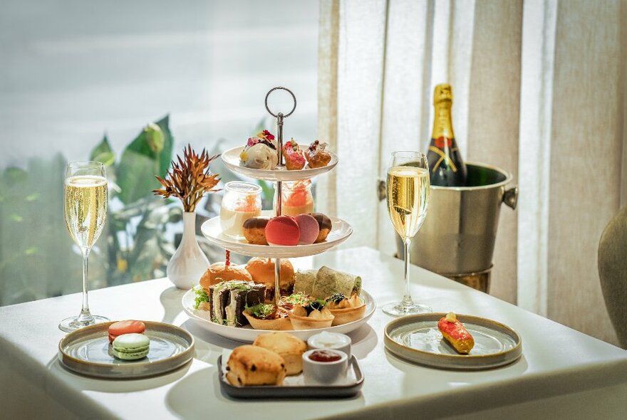 A table set for a high tea with a three-tiered stand of sweet and savoury treats, glasses of sparkling wine and and an ice bucket containing a bottle of wine.
