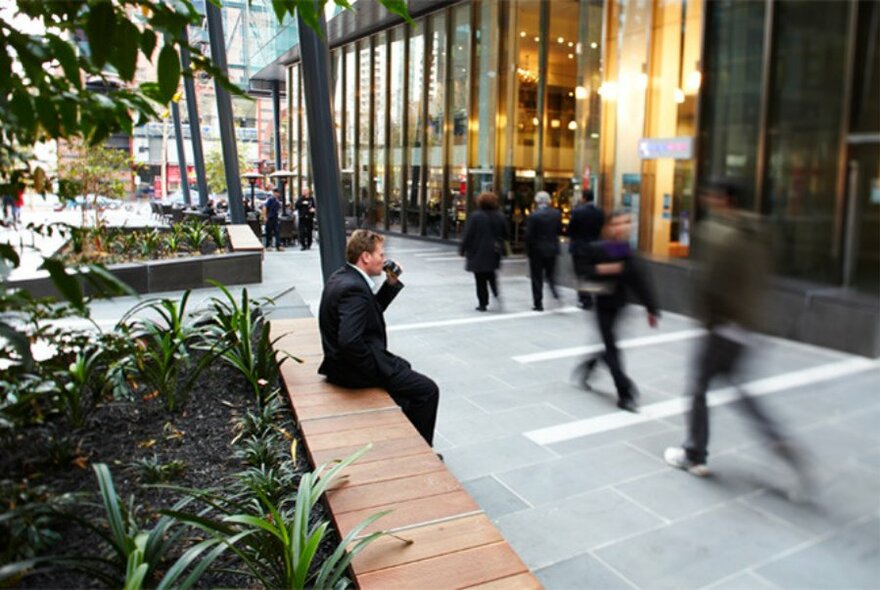 Pedestrian walkway with glass windows, people in motion, and a man sipping a coffee sitting on side of a planter box.