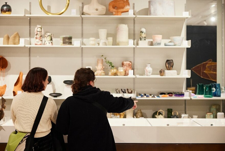 People looking at craft objects displayed on white shelves against a wall in a retail space.