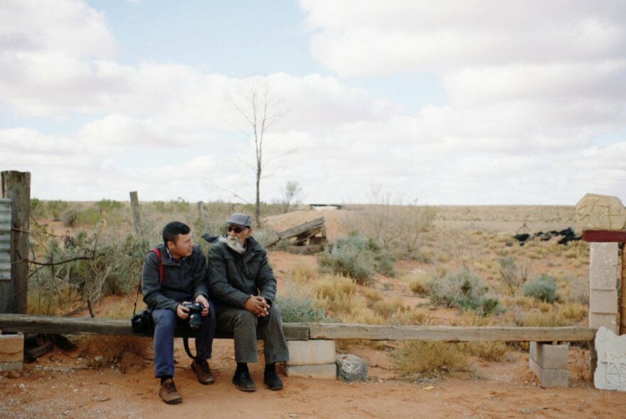 Two people sitting next to each other on a makeshift simple bench in an outback landscape setting.