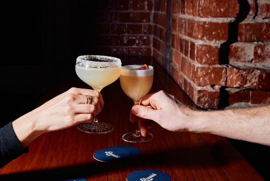 Two hands holding cocktail glasses, clinking them together as though in a 'cheers'.