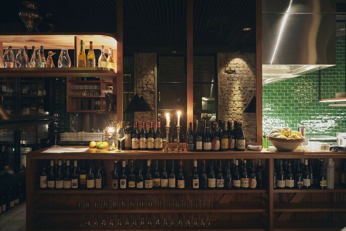 Dramatically lit bar with bottles lined up, green-tiled kitchen area to the right.