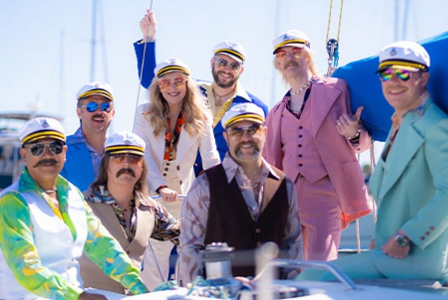The members of the band Yacht Rock Revival, on a yacht and wearing pastel-hued waistcoats and jackets with captains hats.