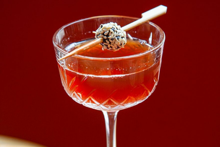 A reddish coloured cocktail in a small crystal glass, with a garnish resting on its edge.