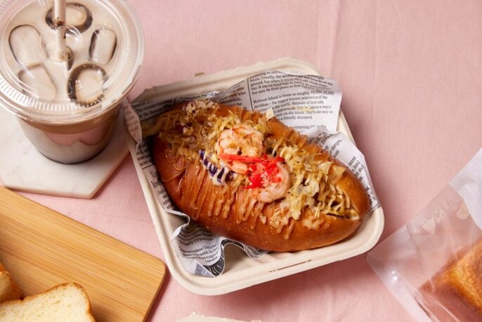 Filled bread roll in a takeaway container with an iced beverage in a plastic cup alongside.