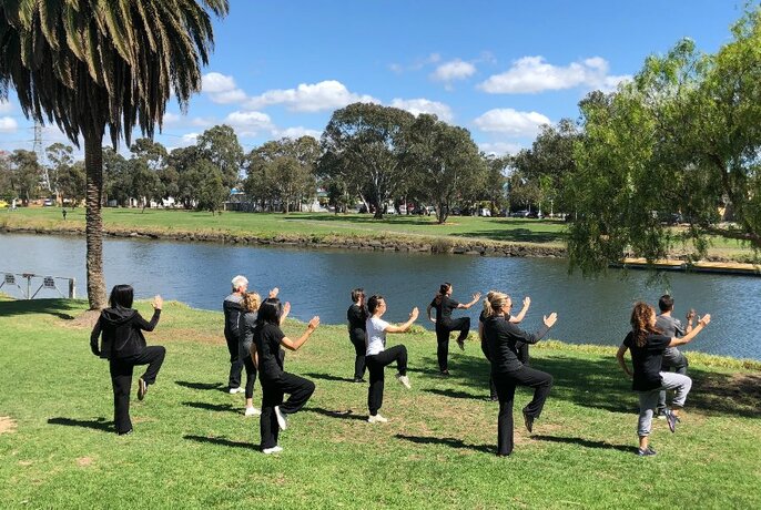 Group of people standing on grass outdoors, near a river bank, holding standing yoga poses.