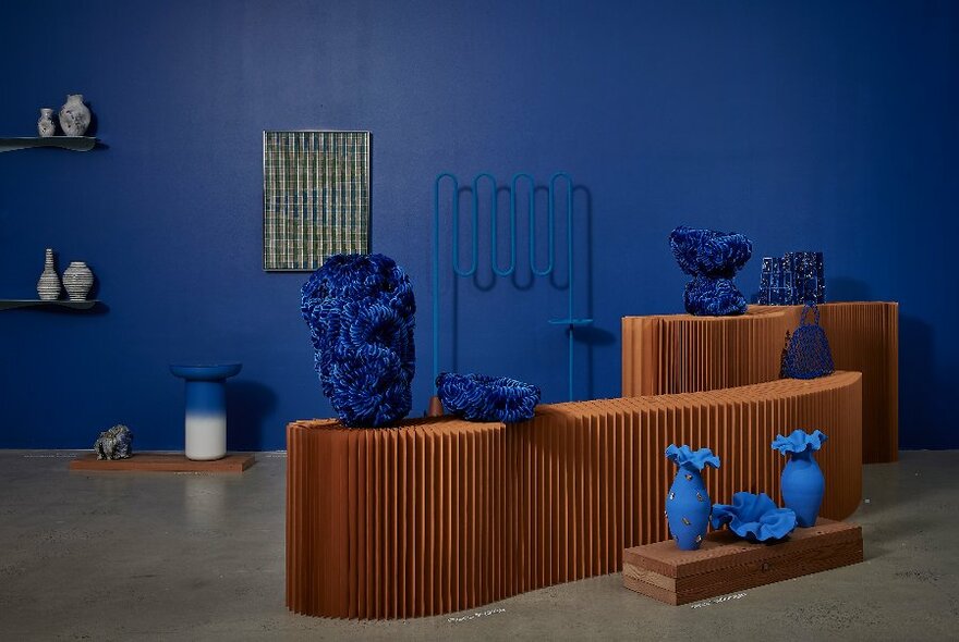 Objects and works of art and craft displayed on wooden display cases and hanging on the blue walls of a gallery showroom.