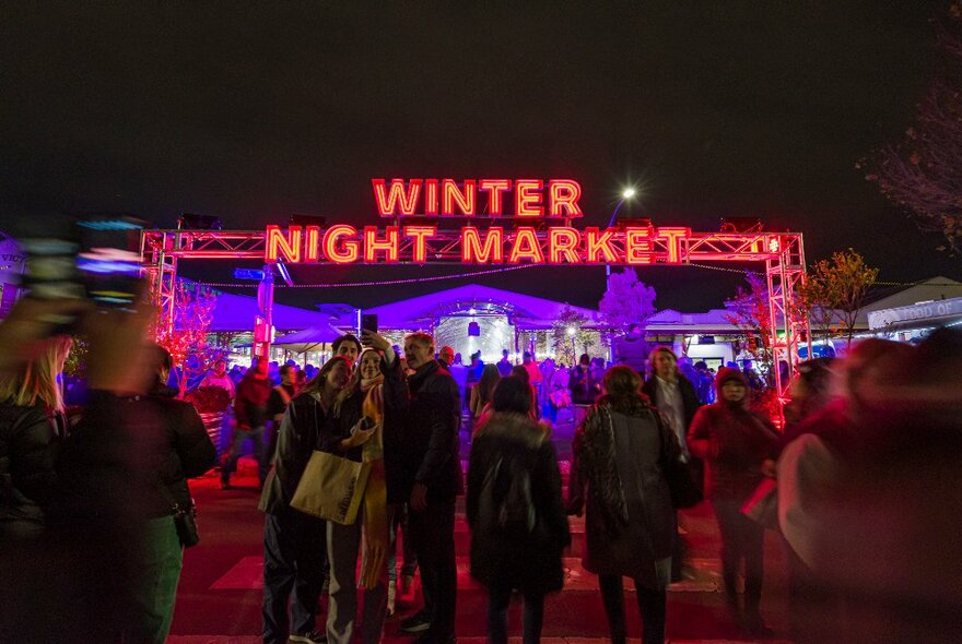 People walking towards a red illuminated archway that reads Winter Night Market. 