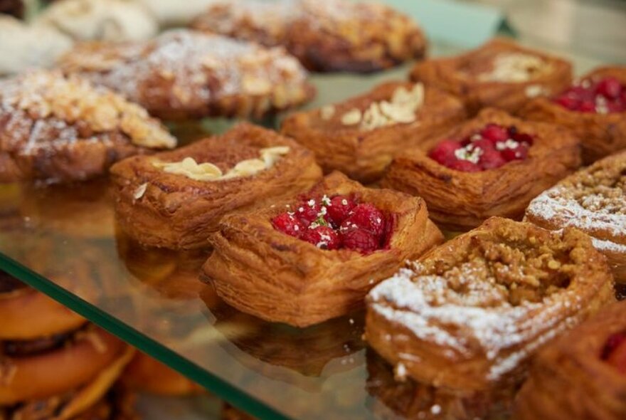 Close up shot of pastries with raspberries, almonds, and powdered sugar on them, placed on a glass shelf.