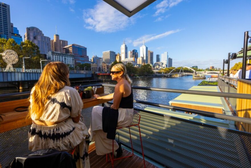 Two women sat at bar bench on stools facing the Yarra River. Blue sky and skyscrapers in background.