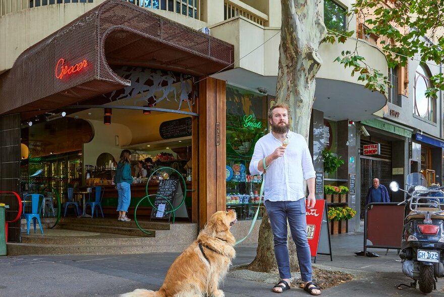 A man eating an ice cream in front of a grocer while walking a golden retriever on a leash.