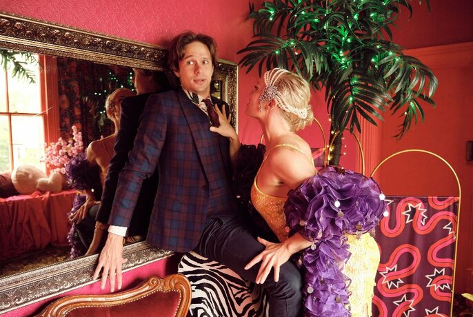 Man pushed up against large, gilt mirror by woman in strappy white dress and purple feather boa.