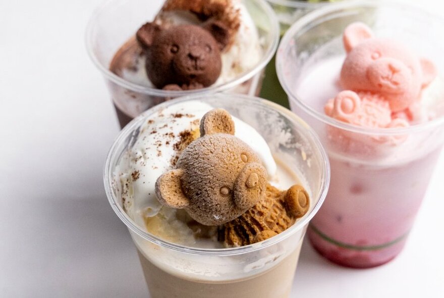 Three take-away plastic drink containers filled with a milky beverage and topped with a bear-shaped biscuit.