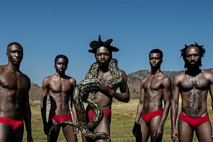 Five men wearing only red underpants staring at the viewer while posed outdoors in a desert landscape against a blue sky; the central figure has a large python snake coiled around his neck and naked torso.