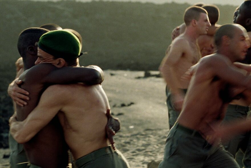 Movie still of bare-chested military members embracing.