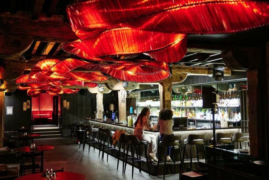 Two women sitting at the bar at an underground restaurant with a red sculpture ribbon light overhead.