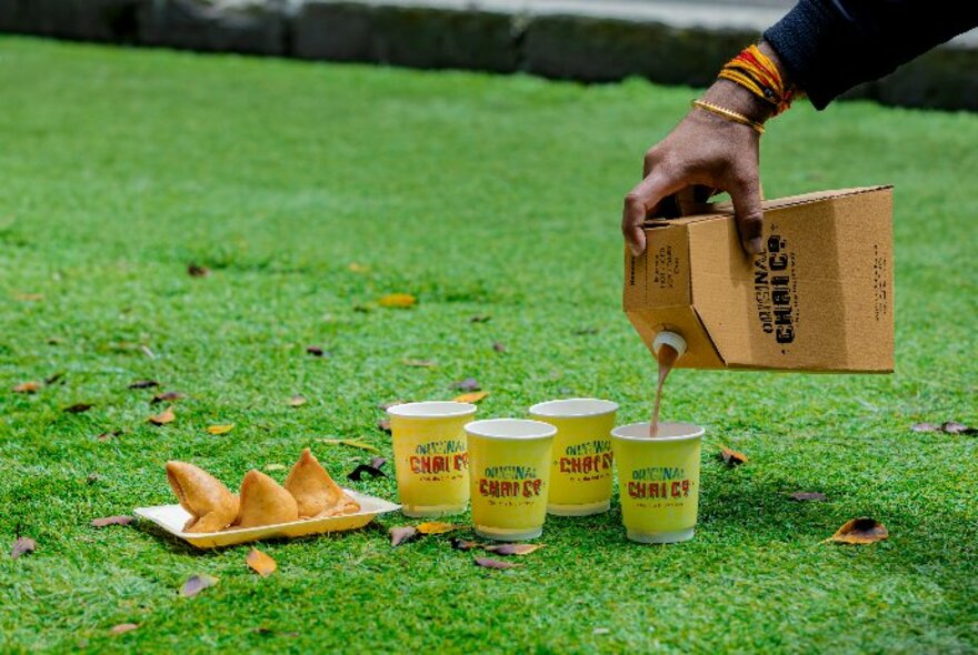 Chai being poured from a cardboard flask into takeaway cups, resting on grass in a park, with a plate of samosas alongside.