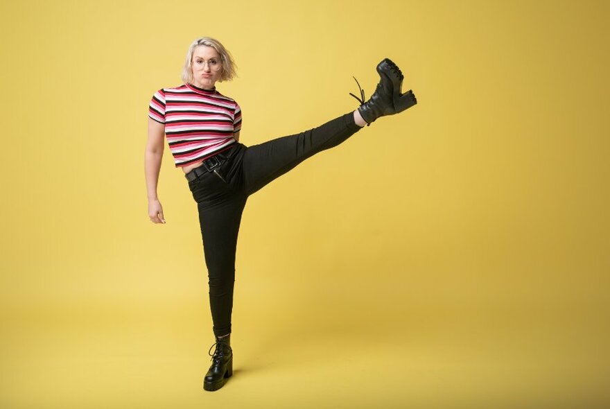 Person standing on one leg, the other raised high in the air to the side, wearing striped T-shirt against yellow background.