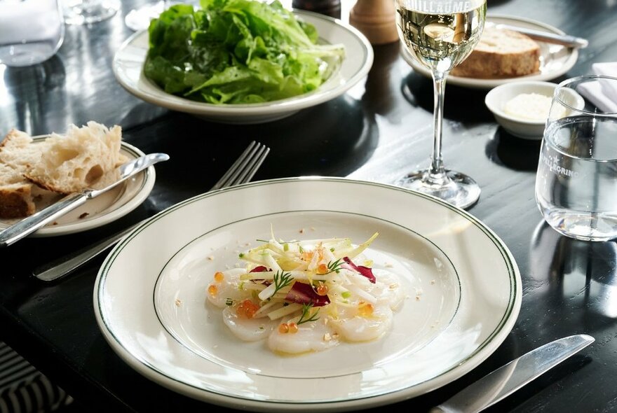 A white plate of Abrolhos Island scallop on a dark table with cutlery, wine and water glasses, a bread plate and a loose-leaf salad.