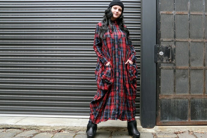 Woman in long, loose flowing red tartan dress and black boots and beanie in front of a steely grey garage door.