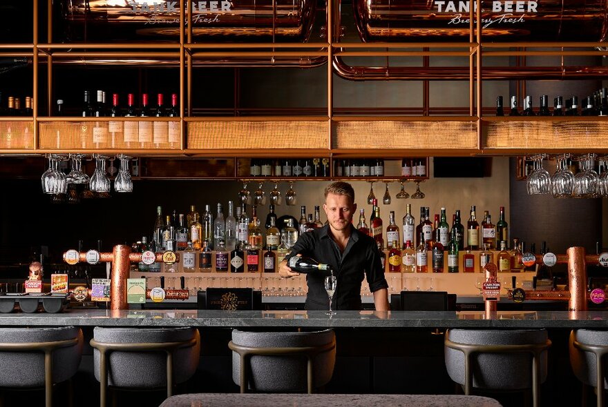 Bar tender pouring wine from a bottle at a bar with barstools, stone bench top, rear display of bottles and overhanging wine shelf.