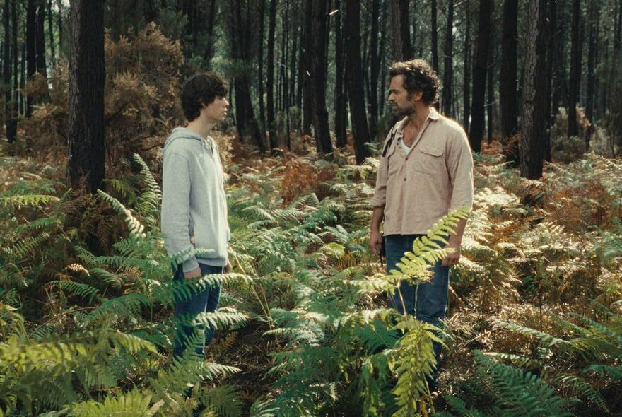 Two men facing each other outdoors in a forest setting among fern bushes; a still from a movie.