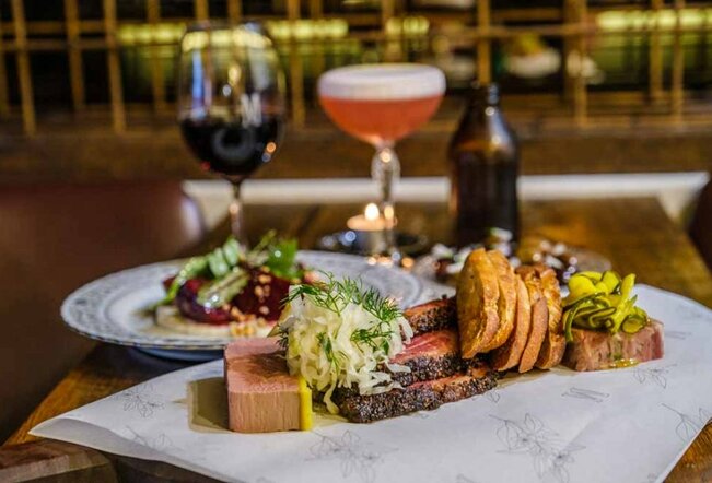 Charred meat, crudites and other cheese board items on a plate in front of a cocktail