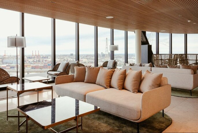 Long apricot-coloured lounges in middle of room with coffee tables, lamps and armchairs, and floor-to-ceiling windows.