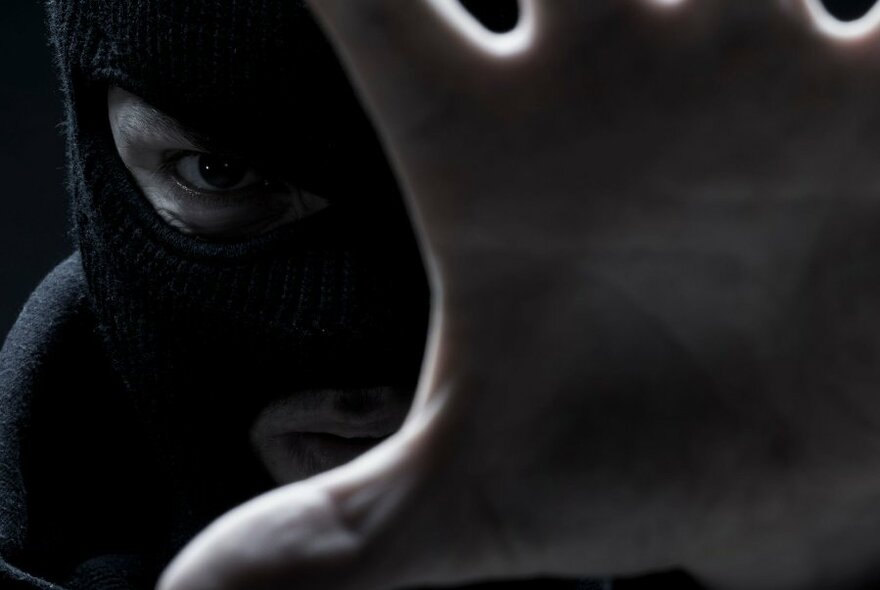 A dark image of a person wearing a balaclava with their hand up to the camera. 