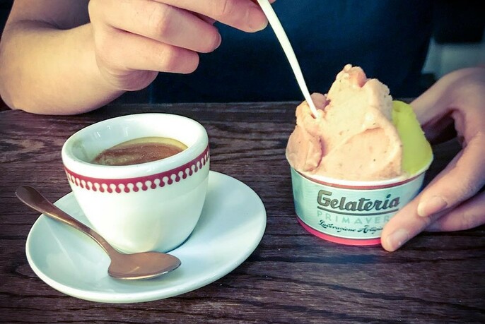 Wooden counter with expresso in a cup and saucer with spoon, and hand dipping a plastic spoon into a takeaway gelati cup.