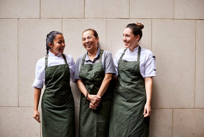 Three smiling and laughing women in chef white shirts and dark khaki aprons, standing in front of a light coloured wall.