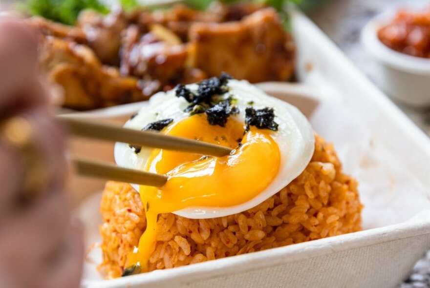 Chopsticks plunging into a lightly fried egg perched on top of a mound of rice in a white dish.