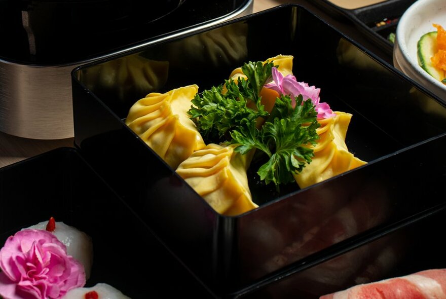 Four dumplings or wontons arranged in a square black dish, with a parsley garnish.