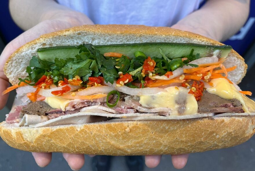 A single Vietnamese Banh Mi roll being held in the palms of a person, showing the delicious fillings including cucumer, coriander, roast pork and chilli.