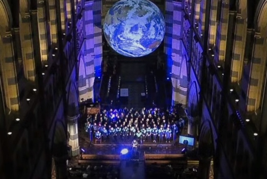 Large choir of people on the altar area of a cathedral, view looking down, columns on either side, with a large blue image of the earth displayed above the singers.