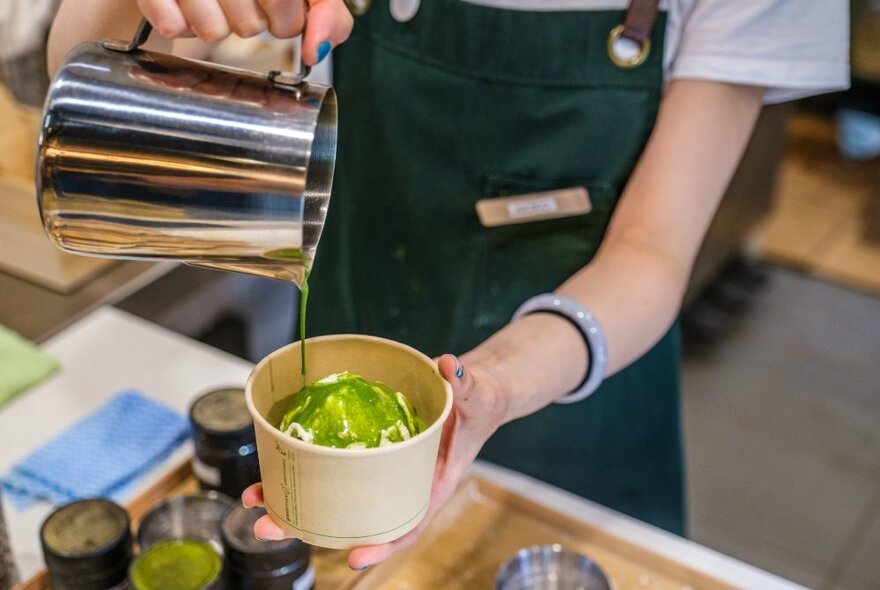 Person working behind a food counter pouring green matcha liquid topping over ice cream in a takeaway cardboard container they hold in their hand. 
