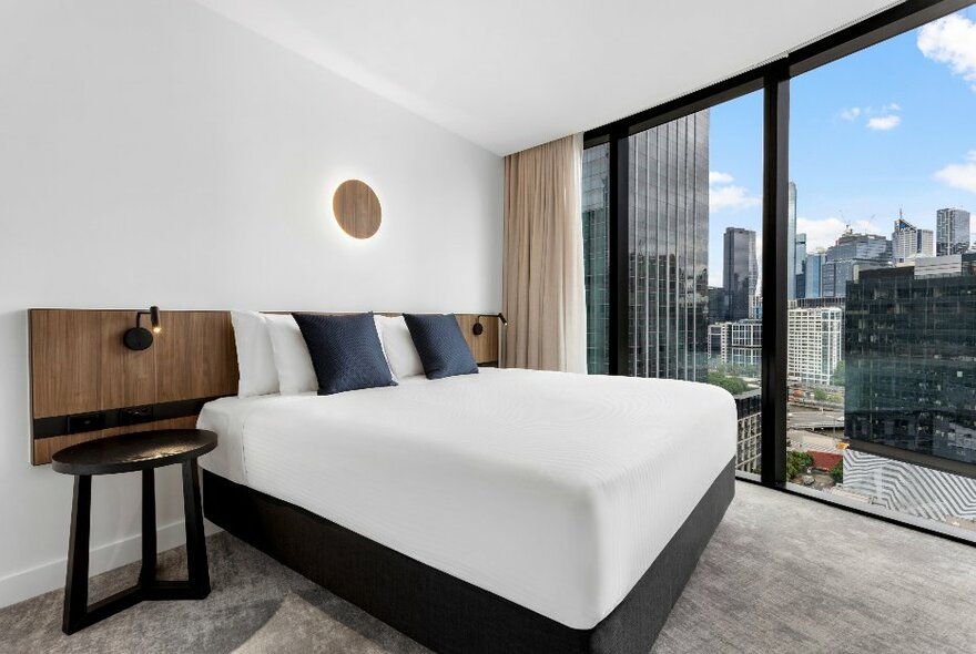 Hotel room with white linen on bed, grey carpet and floor to ceiling view out onto city buildings.