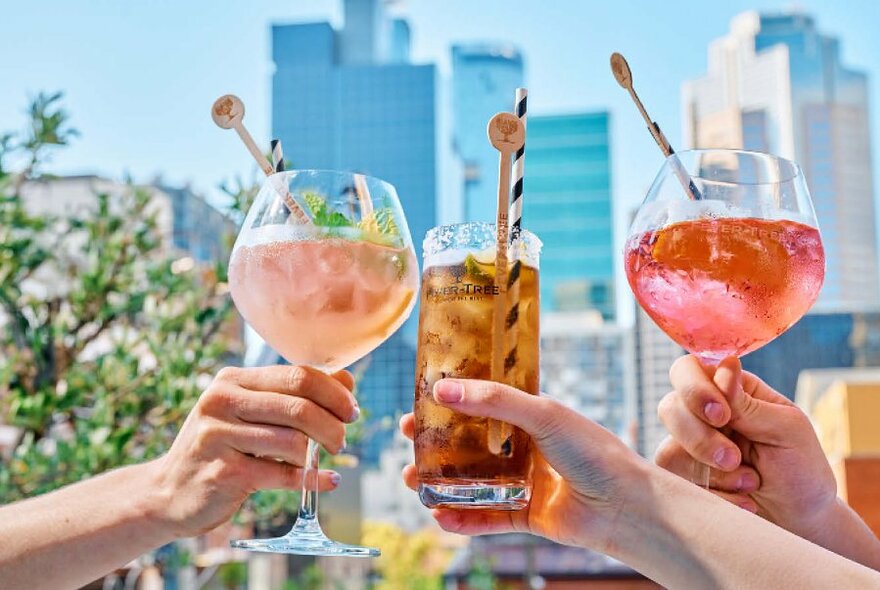 Three different cocktails being held up to the city skyline.