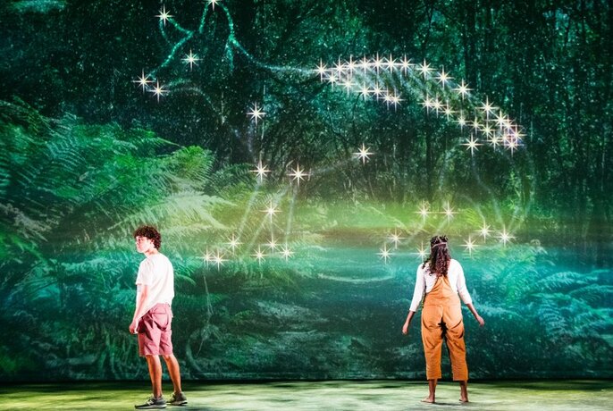 Tow performers/actors with their back to the audience, standing on a stage, a large green rainforest mural in the background with an outline of a Tasmanian Tiger on it.