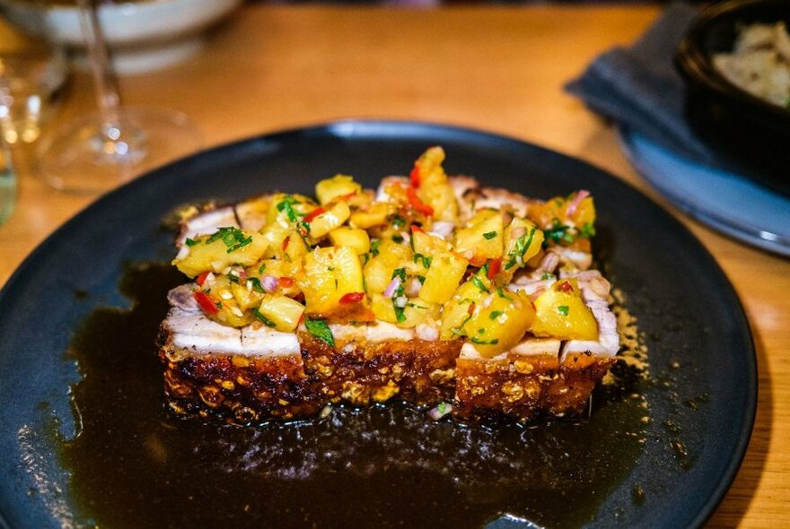 Grilled meat dish, topped with a sweetcorn salsa, served on a dark plate.