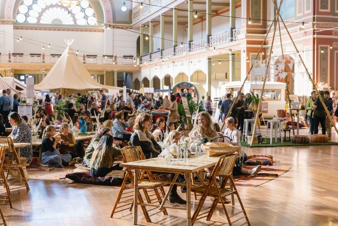 Interior shot of exhibition hall with people seated at table and chairs and on the floor on cushions, with tee-pee and yurt structure behind, as well as fairy lights above.