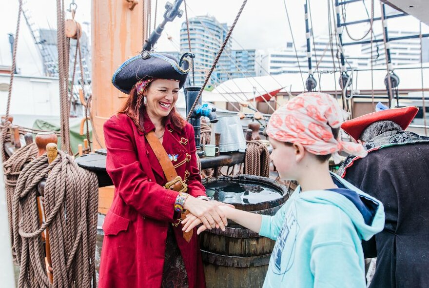 A person dressed as a pirate in a red coat shaking hands with a child wearing a head scarf aboard an old sailing ship.