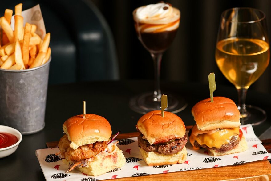 Three sliders on a wooden tray, fries, a dessert and a drink on a table.