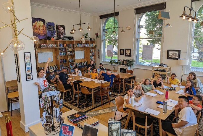 Large studio space with young children and teenagers sitting around at tables, smiling and writing on paper, the walls of the space covered in shelves of books.