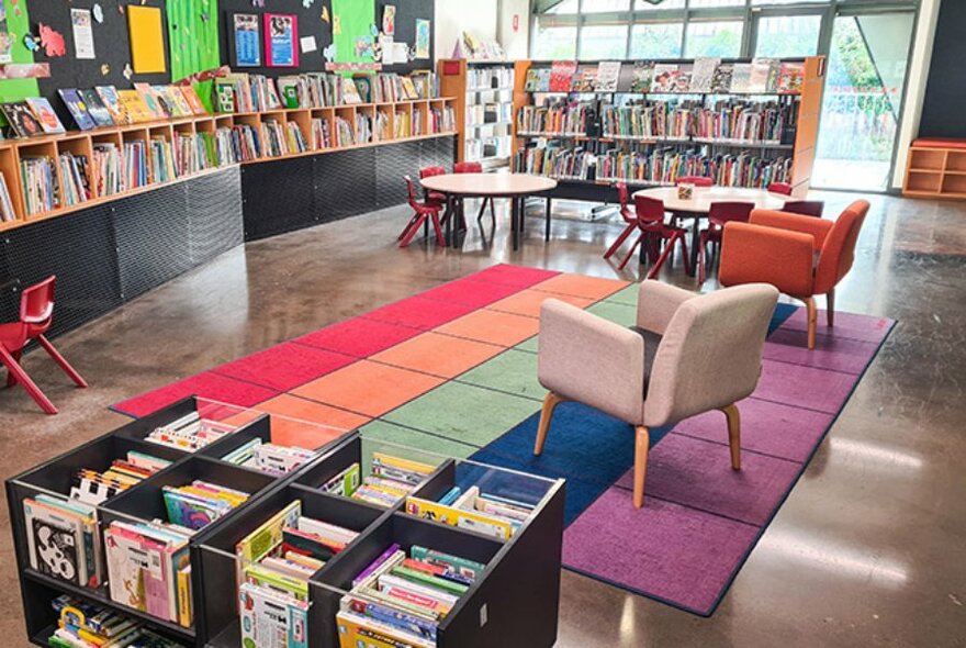 Interior of East Melbourne Library with shelves of books, a rug featuring coloured squares on the floor and armchairs.