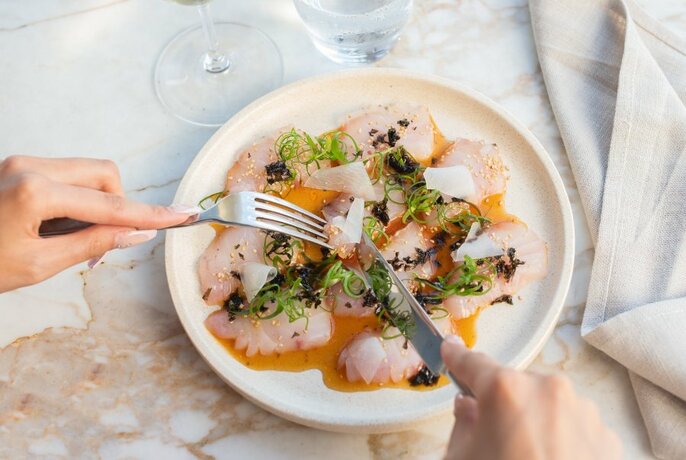 Hands holding cutlery slicing into dish of raw fish  with amber coloured sauce and micro herbs.