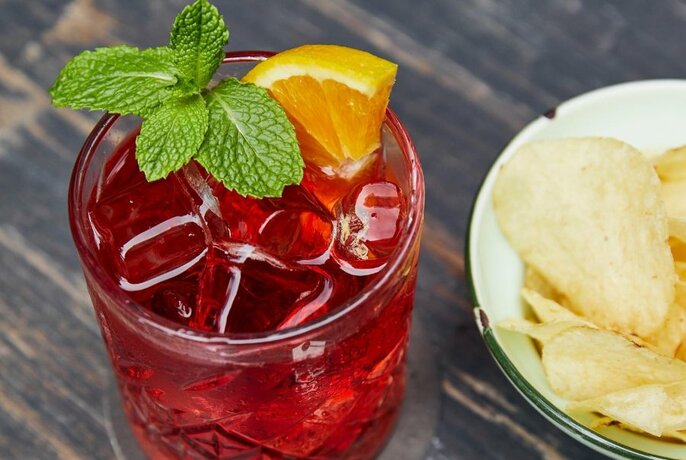 Red cocktail in a short glass, garnished with mint and an orange slice, next to a bowl of crisps.